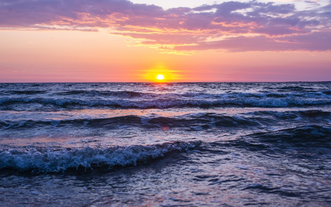 A sun shining during golden hour with pink and purple cloud colors and sea waves