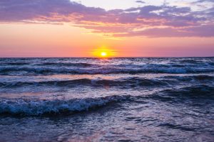 A sun shining during golden hour with pink and purple cloud colors and sea waves